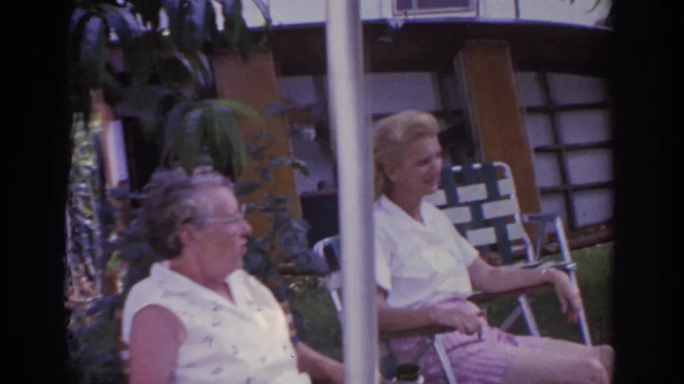 Two seated women appear in a 1963 New Jersey yard or porch view, partly framed by posts and foliage.
