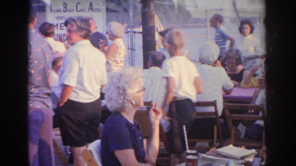 A crowd of adults sits and stands around tables in New Jersey home-movie footage, with a hand-lettered sign partly visible.