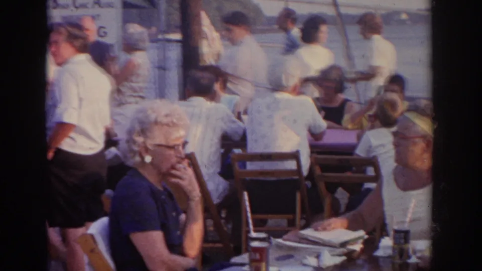Adults sit and stand around tables in a 1963 New Jersey outdoor gathering, with faces and figures partly obscured by the 8mm frame.