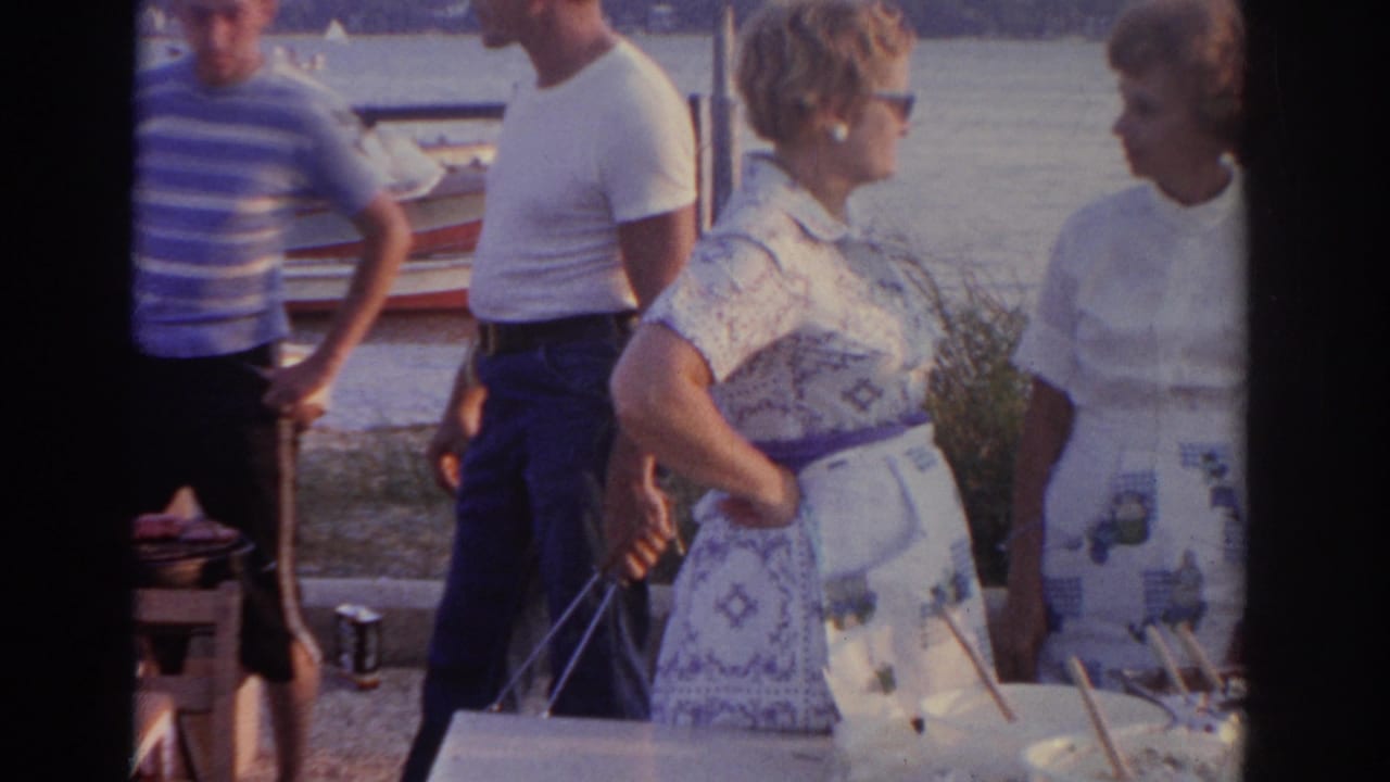 Adults stand around an outdoor table near water and small boats.