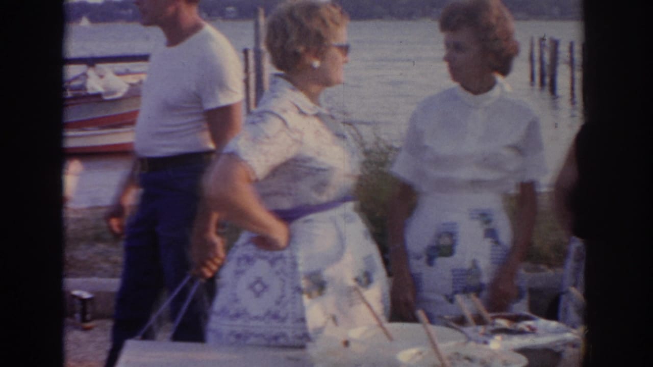 Two women stand beside a table as a man and boats appear behind them.
