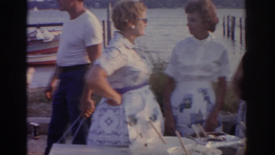 Two women in light summer clothing stand at a waterfront table in 1963 New Jersey, with a man and dock area behind them.