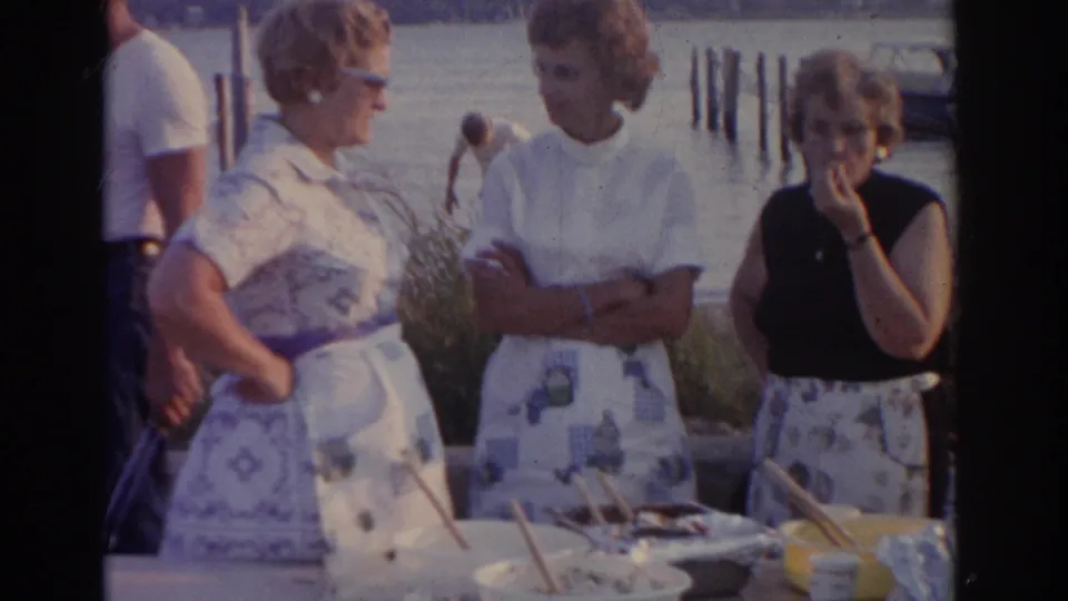 Three women gather around a waterfront table in a 1963 New Jersey home movie, with dishes, cups, and dock pilings visible.