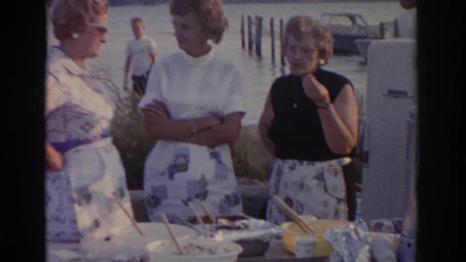 Women in 1960s clothing stand by a waterfront meal table in New Jersey, with dock pilings and a small boat in the background.