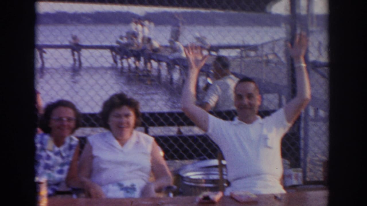 Adults seated at a waterfront table as a man in a white shirt waves behind a chain-link fence.