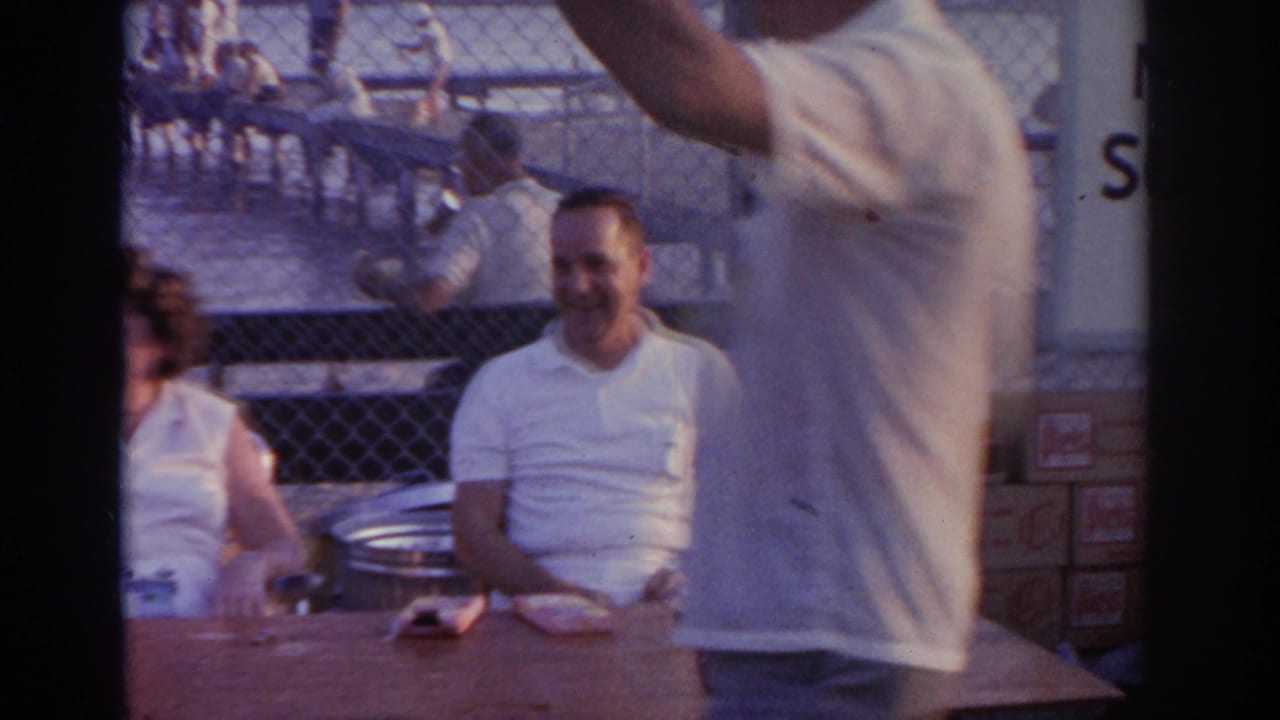 Smiling man seated at a table while another person stands close to the camera near the waterfront.