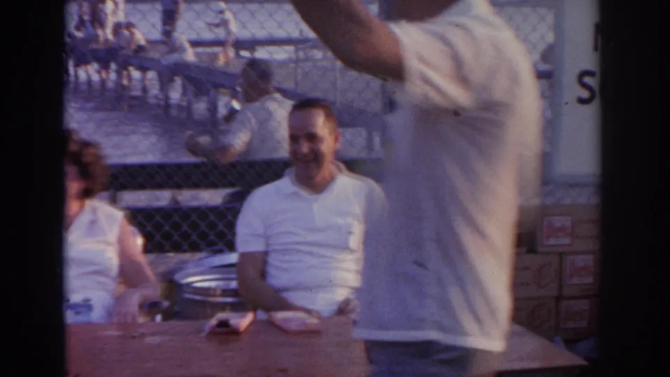 1963 New Jersey waterfront frame with a seated smiling man, nearby adults, chain-link fencing, and people gathered on a pier over the water.