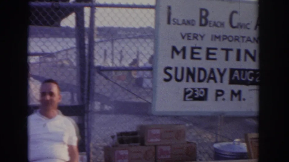 1963 New Jersey home-movie frame showing an Island Beach Civic Association meeting sign near chain-link fencing and waterfront activity.