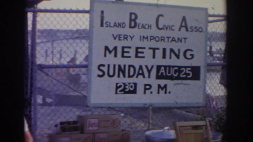 1963 New Jersey frame focused on an Island Beach Civic Association meeting sign announcing a Sunday 2:30 p.m. meeting near the water.