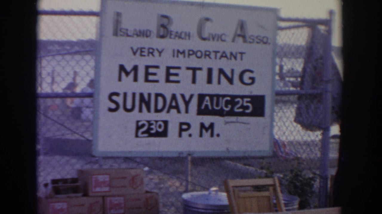1963 New Jersey 8mm frame showing an Island Beach Civic Association meeting sign posted beside fencing near docks and water.