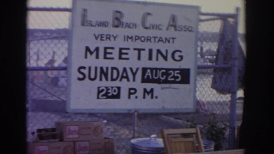 1963 New Jersey 8mm frame showing an Island Beach Civic Association meeting sign posted beside fencing near docks and water.