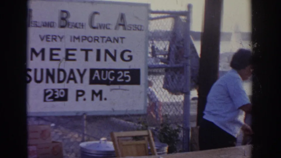 1963 New Jersey 8mm frame with the civic meeting sign in front of a waterfront fence and a woman partly visible nearby.