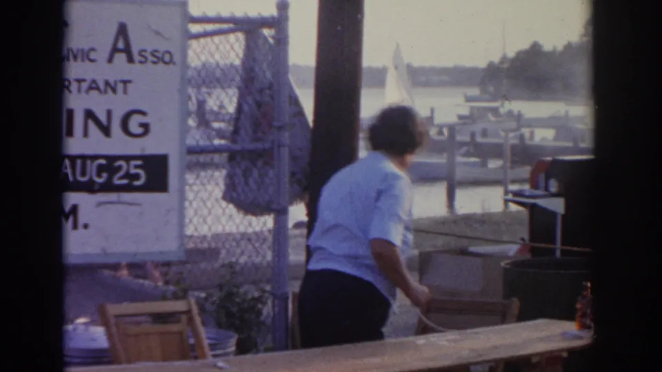 1963 New Jersey dockside frame showing a woman by a cooking or serving area, with boats and water behind her.