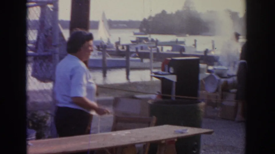 1963 New Jersey waterfront home-movie frame showing a woman near a grill or food setup with boats and docks behind her.