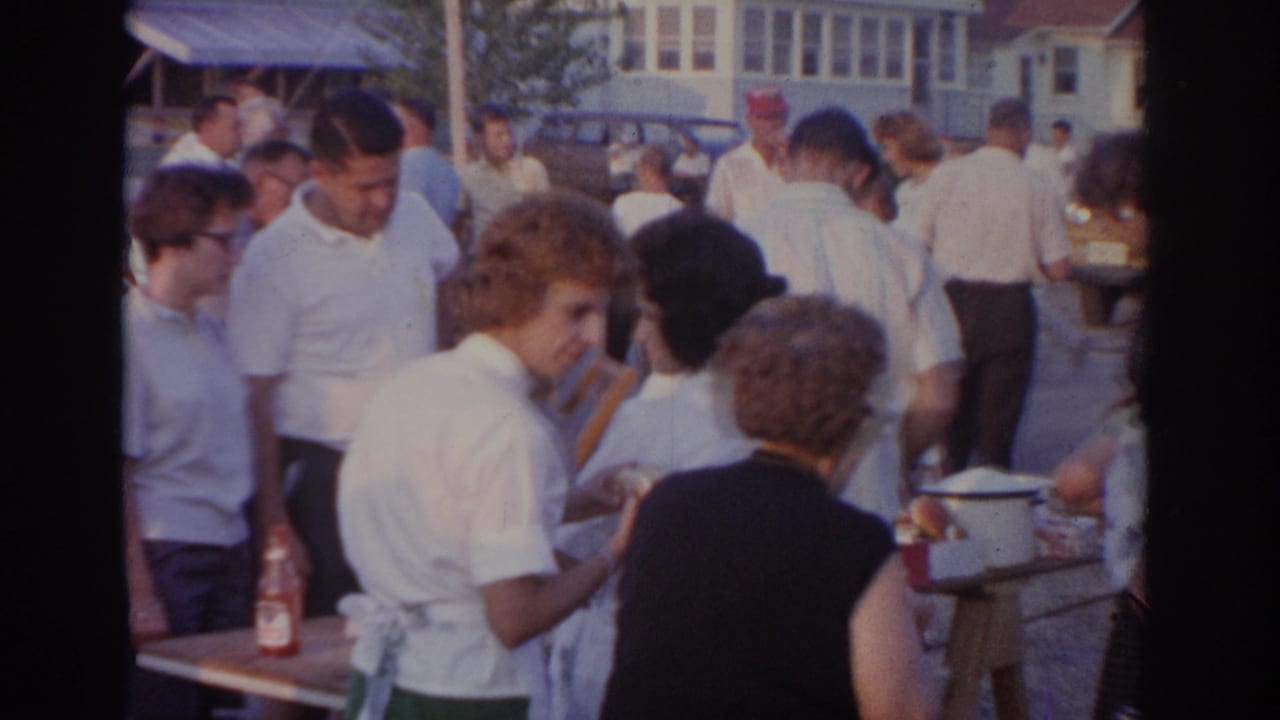 Adults gather around outdoor serving tables at a cookout with houses and cars behind them.