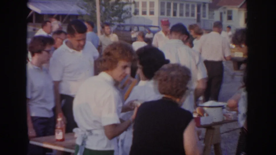 1963 New Jersey 8mm frame showing adults in summer clothing near food tables at an outdoor cookout, with houses and parked cars in the background.