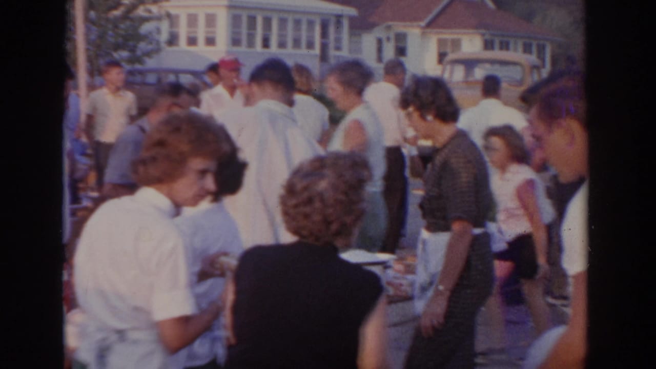 Crowd of adults stands around food tables at an outdoor cookout near parked cars.