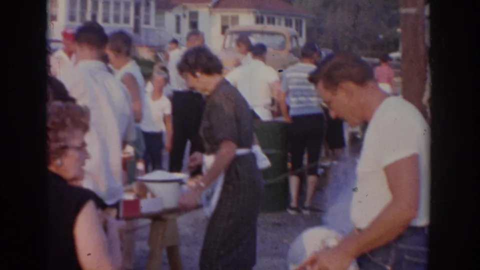 1963 New Jersey home-movie frame with a man at a grill and adults gathered around serving tables during an outdoor cookout.