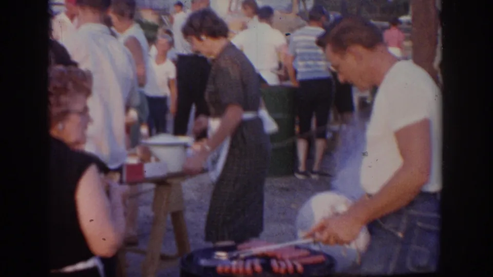 Close 8mm view from a 1963 New Jersey cookout, showing a man grilling hot dogs or sausages beside guests and serving tables.