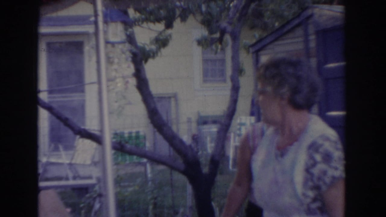Older woman in a light dress stands near a tree and house exterior.
