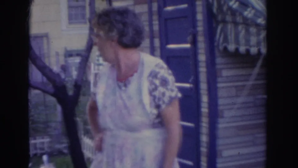 A woman in a light dress looks across a New Jersey yard, with a blue door, house siding, tree, and fence visible.