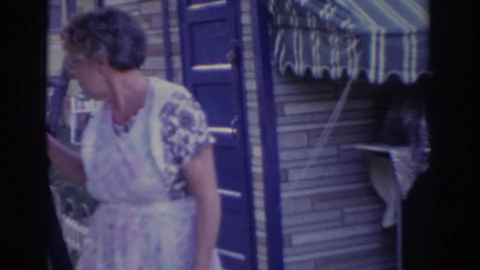 The woman gestures near the blue door and striped awning, with another person partially visible at the edge of the frame.