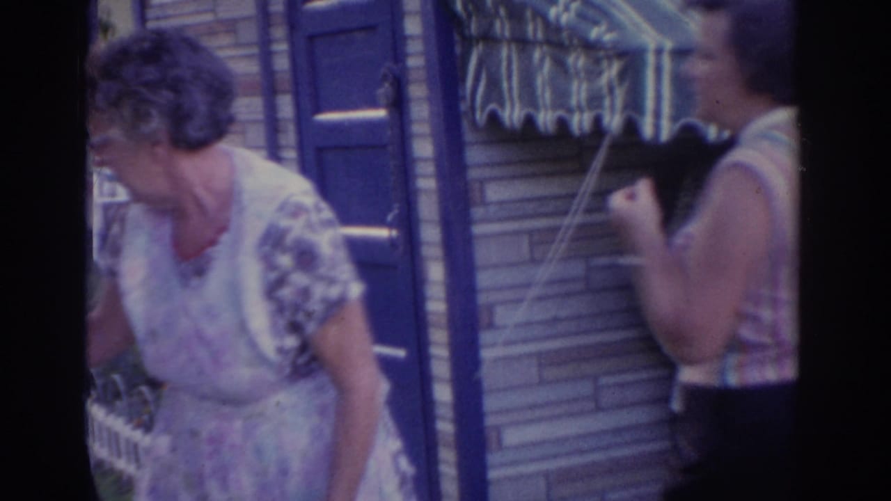 1963 New Jersey home-movie frame showing two adult women outside a brick house, one in a floral dress and one near a small grill.