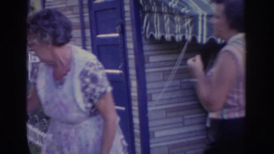 1963 New Jersey home-movie frame showing two adult women outside a brick house, one in a floral dress and one near a small grill.