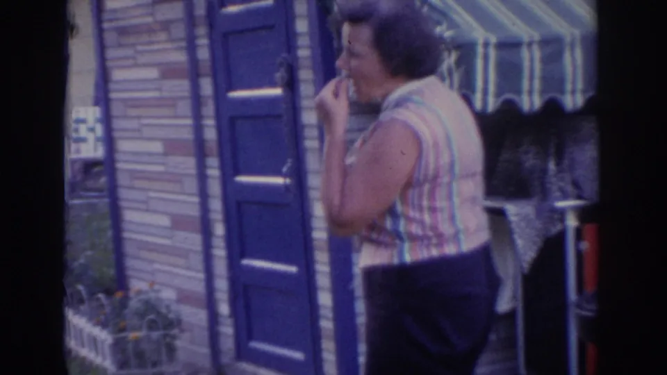 1963 New Jersey frame of an adult woman in a striped sleeveless top appearing to taste food beside a brick house and small grill area.
