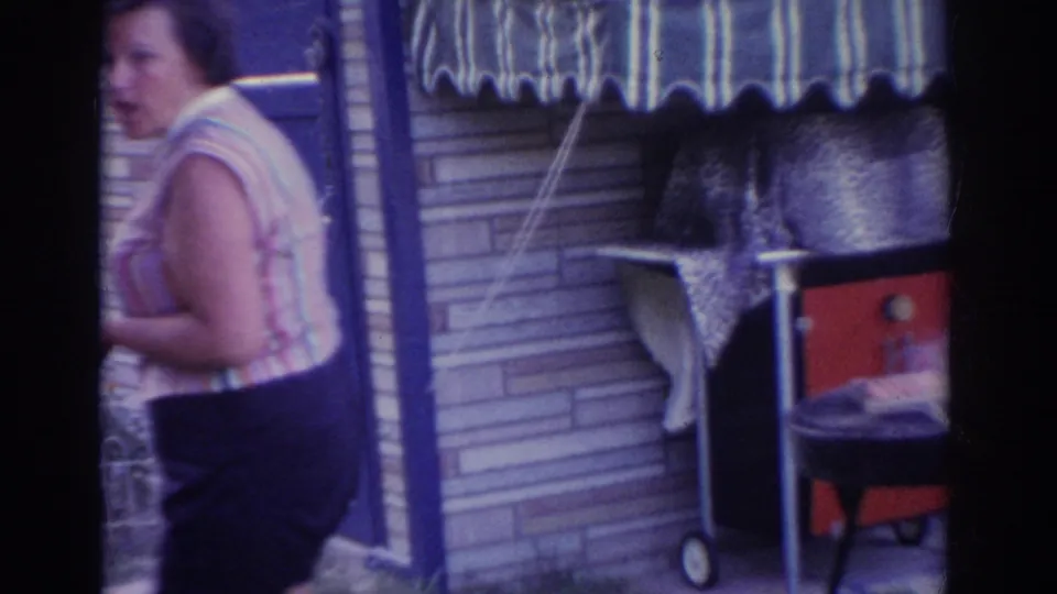 1963 New Jersey home-movie frame showing an adult woman passing a brick house exterior with striped awning and red grill nearby.