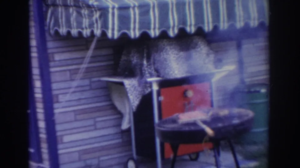 1963 New Jersey home-movie frame with smoke rising from an outdoor grill under a blue and white striped awning, near a red cooler and green trash can.