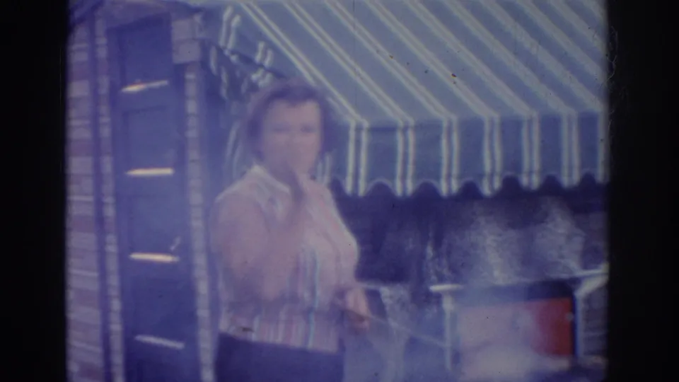 1963 New Jersey home-movie frame showing an adult woman in a striped sleeveless shirt near a smoking backyard grill under a striped awning.