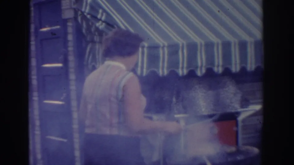 1963 New Jersey home-movie frame showing an adult woman from the side or back as she tends a smoking outdoor grill under a striped awning.