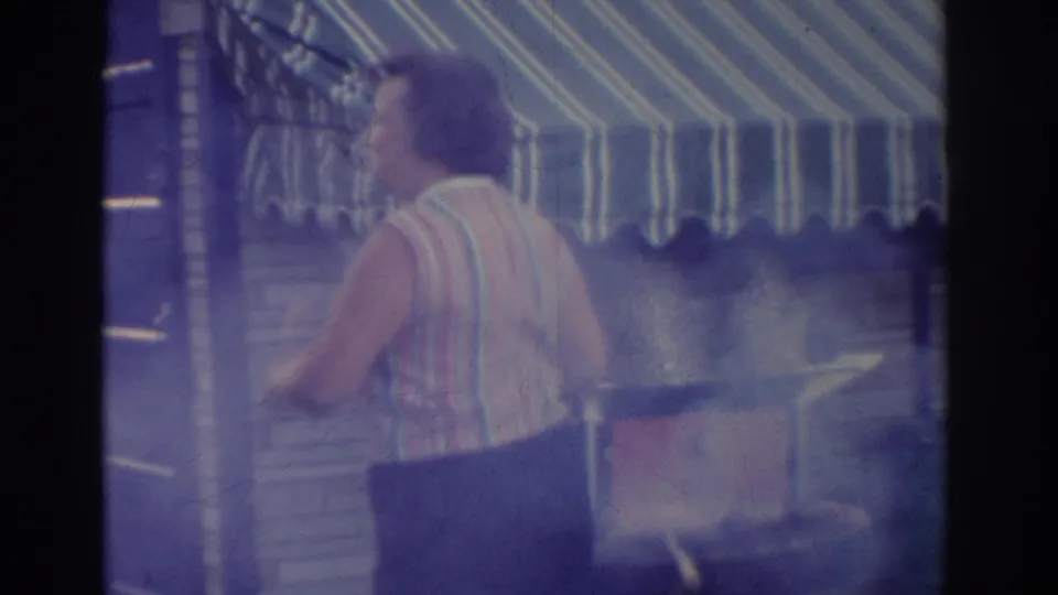 A rear view shows the woman continuing to move near the smoking grill beneath the striped awning in the New Jersey home movie.