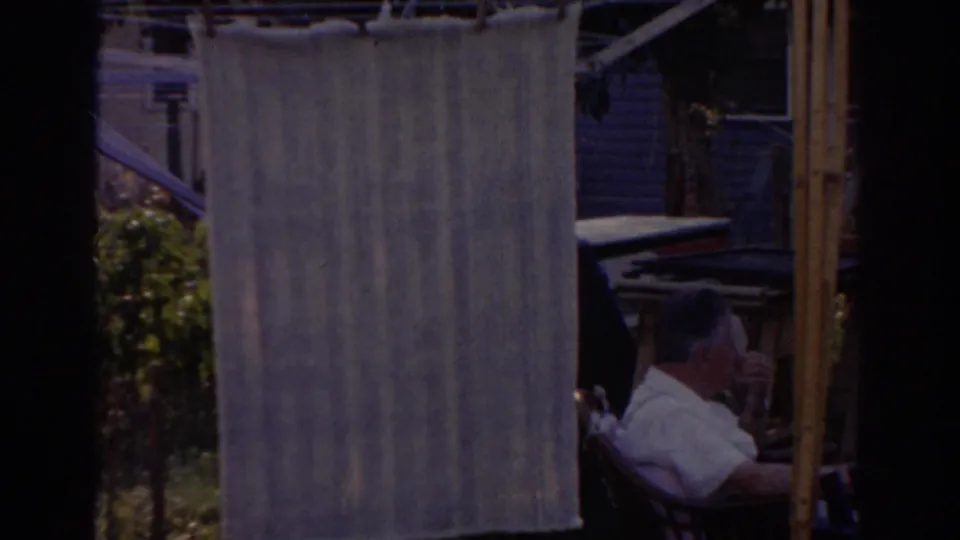 In a 1963 New Jersey backyard, a man sits in a chair near a hanging cloth and house exterior.