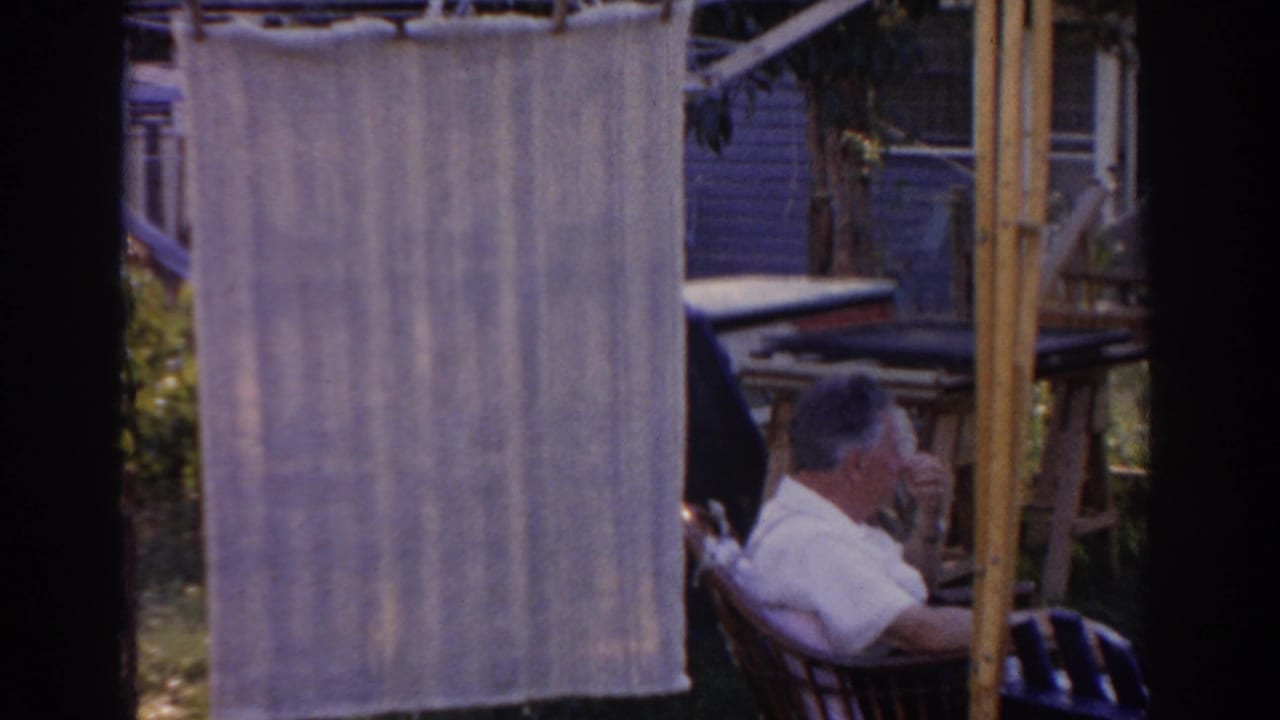 Man in a white shirt sits in a yard chair near hanging fabric and outdoor furniture.
