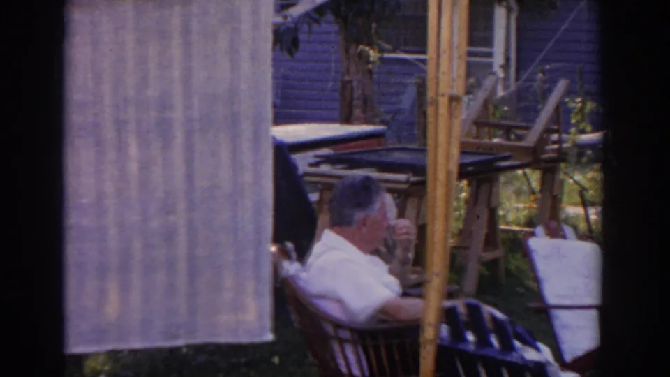 A seated man in a 1963 New Jersey yard appears to drink or rest beside hanging fabric and furniture.