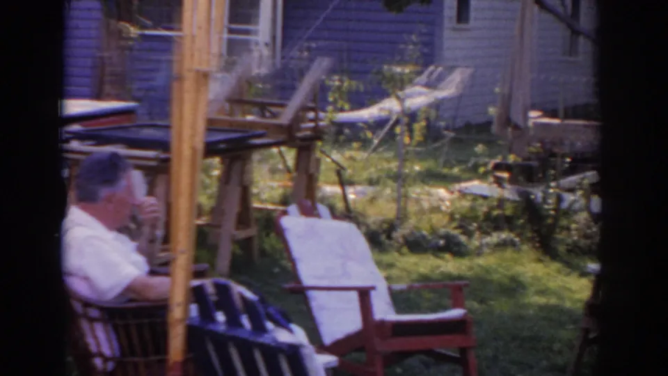The man remains seated in the shaded yard, with a hammock, chairs, and house siding visible behind him.