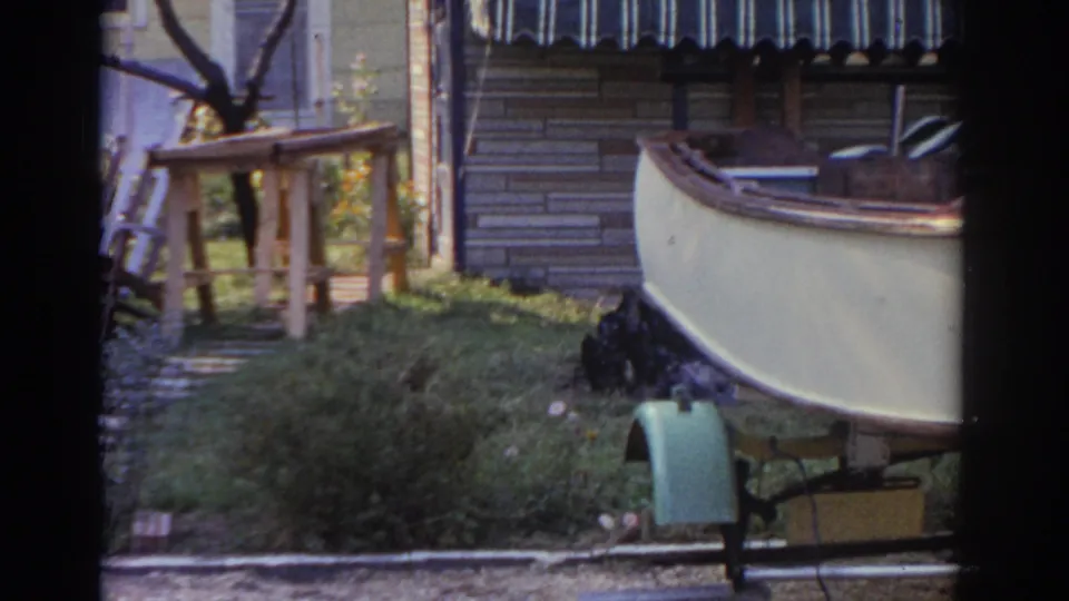 As the view shifts, a wooden table or stand and outdoor chairs appear near the boat trailer in the New Jersey backyard.