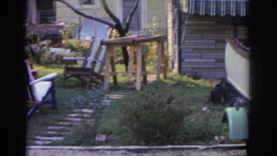 The final frame shows lawn chairs, a stone path, grass, and a wooden stand beside the edge of the boat trailer near the house.