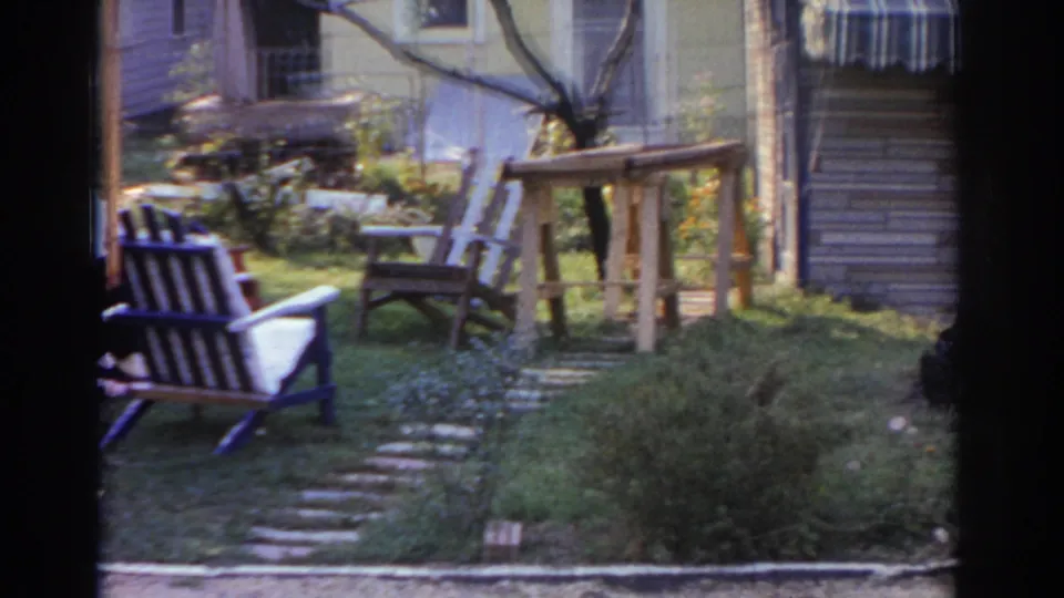 1963 New Jersey home-movie frame showing a suburban backyard with wooden chairs, a workbench, garden path, lawn, and house exterior.