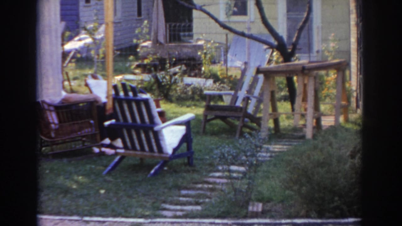 Backyard view with wooden chairs, a wicker chair, workbench, path, and house exterior.