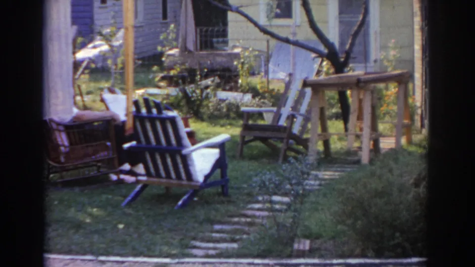1963 New Jersey backyard view with lawn chairs, a wicker seat, workbench, stone path, shrubs, and part of a house visible.