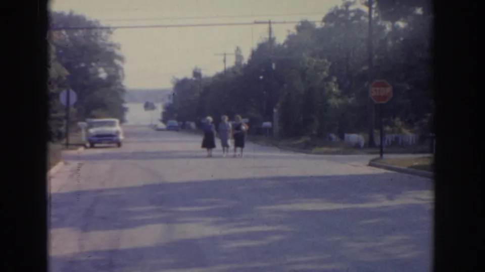 1963 New Jersey home-movie frame of women walking down a tree-lined residential street near a stop sign and period car.