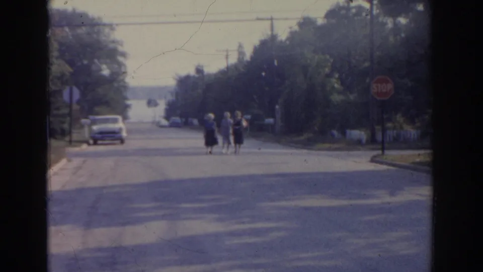 Three women in dress clothes walk along a New Jersey suburban street in 1963, with a stop sign, parked car, trees, and utility lines visible.