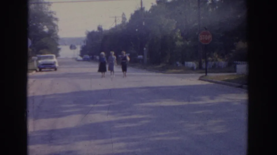 Three women dressed for an outing approach the camera on a quiet New Jersey residential street, with a stop sign, trees, and parked vehicles behind them.
