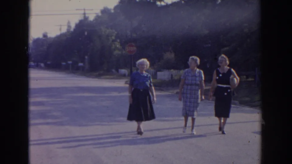 Three adult women in 1960s dresses and hats walk together toward the camera on a New Jersey street, with a stop sign and trees in the background.