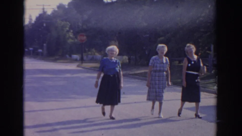 Three older women in dresses and skirts walk together across a bright New Jersey street, with trees and a stop sign in the background.