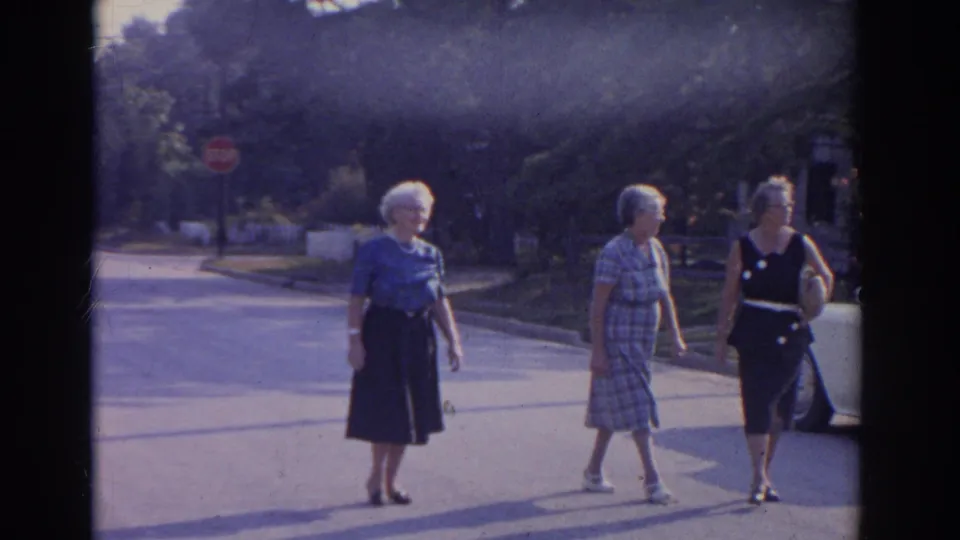 Three older women walk past the camera on a sunny New Jersey street, with a partial parked car entering the right edge.