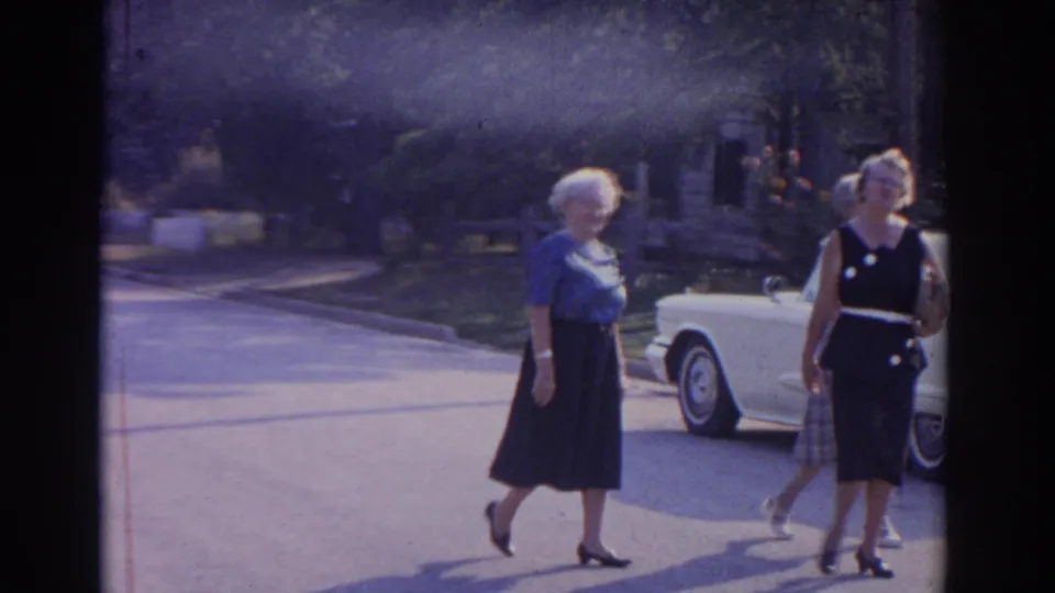 1963 New Jersey home-movie frame showing three older women walking past a parked white classic car on a residential street.